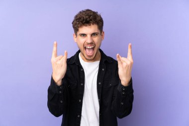 Caucasian man isolated on purple background making horn gesture