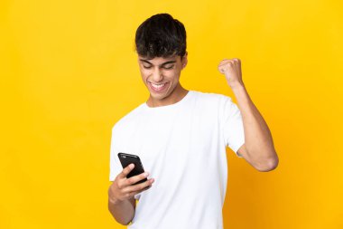 Young man over isolated yellow background using mobile phone and doing victory gesture