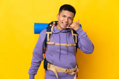 Sarı arka planda izole edilmiş büyük bir sırt çantası olan genç dağcı telefon hareketlerinde bulundu. Bana geri dönüş işareti ver.