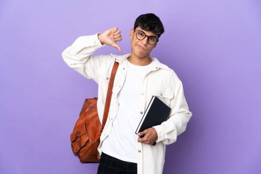 İzole edilmiş mor arka planda genç bir öğrenci, olumsuz ifadeyle baş parmağını aşağı gösteriyor.