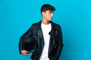 Young man with a motorcycle helmet over isolated blue background looking to the side