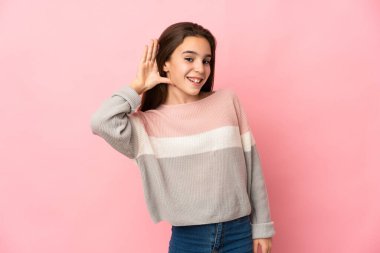 Pembe arka planda izole edilmiş küçük bir kız kulağına elini sürerek bir şeyler dinliyor.