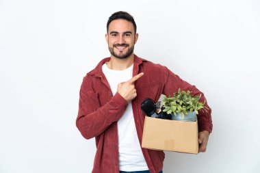 Beyaz arka planda izole edilmiş eşyalarla dolu bir kutuyu alıp bir ürün sunmak için kenarı işaret eden genç beyaz bir adam hamle yapıyor.