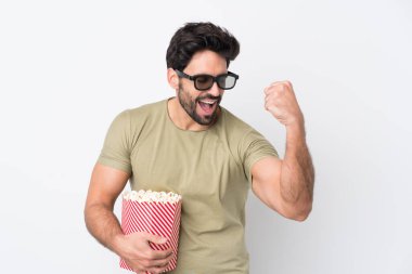 Sakallı, beyaz arka planı olan, 3 boyutlu gözlüklü ve elinde koca bir kova patlamış mısırla yakışıklı bir genç.