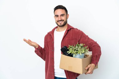 Beyaz arka planda izole edilmiş eşyalarla dolu bir kutuyu kaldırırken beyaz bir adam hamle yapıyor. Davet etmek için ellerini yana uzatıyor.