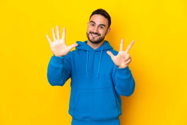 Sarı arka planda izole edilmiş beyaz, yakışıklı genç bir adam sekiz parmaklı sayıyor.