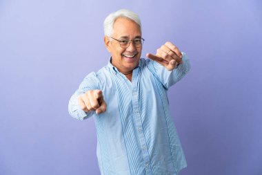 Mor arka planda izole edilmiş orta yaşlı Brezilyalı adam mutlu bir ifadeyle öne işaret ediyor.