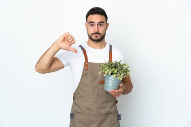 Bahçıvan beyaz arka planda izole edilmiş bir bitki tutuyor. Başparmağını negatif ifadeyle aşağı gösteriyor.