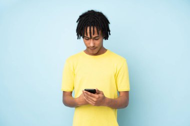 Genç Afro-Amerikan adam mavi arka planda izole edilmiş cep telefonuyla bir mesaj gönderiyor.