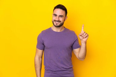 Beyaz tenli yakışıklı adam sarı arka planda izole edilmiş ve en iyinin işareti olarak parmağını kaldırıyor.