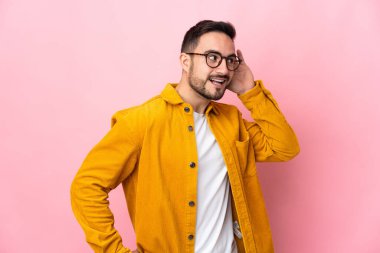 Beyaz tenli yakışıklı adam pembe arka planda izole edilmiş bir şekilde elini kulağına koyarak bir şeyler dinliyor.