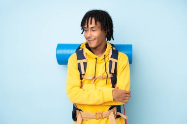 Genç dağcı Afro-Amerikan adam büyük bir sırt çantası ile mavi bir arka planda izole gülüyor