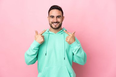 Pembe arka planda izole edilmiş beyaz, yakışıklı bir genç baş parmak hareketi yapıyor.