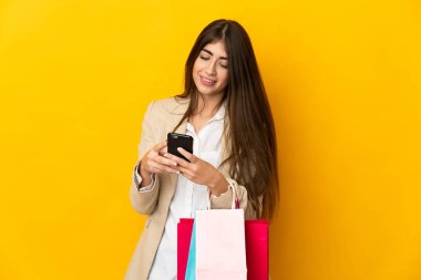 Sarı arka planda izole edilmiş genç beyaz bir kadın alışveriş torbaları tutuyor ve bir arkadaşına cep telefonuyla mesaj yazıyor.