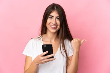 Pembe arka planda izole edilmiş genç beyaz kadın cep telefonu kullanıyor ve yan tarafı işaret ediyor.