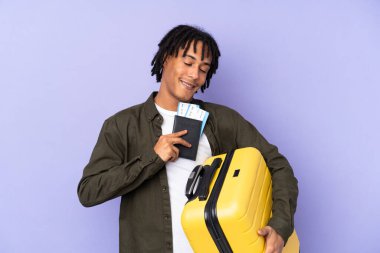 Genç Afro-Amerikalı bir adam mor arka planda izole edilmiş, bavul ve pasaportla tatilde.