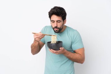 İzole edilmiş beyaz arka planda sakallı genç yakışıklı bir adam elinde çubuklu bir kase erişte tutuyor ve sıcak oldukları için üflüyorlar.