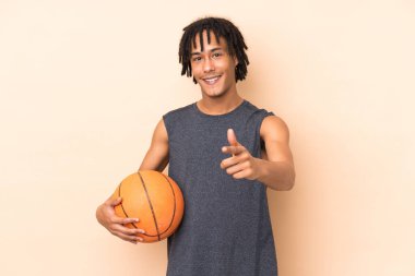 Genç Afrikalı Amerikalı adam bej arka planda izole edilmiş basketbol oynuyor ve ön tarafı işaret ediyor.
