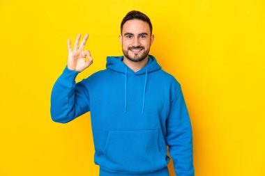 Sarı arka planda izole edilmiş beyaz, yakışıklı genç bir adam parmaklarıyla tamam işareti gösteriyor.