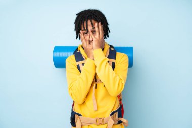 Genç dağcı Afro-Amerikan adam büyük bir sırt çantası ile mavi bir arka planda izole gözleri kapalı ve parmaklarından bakarak