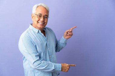 Mor arka planda izole edilmiş orta yaşlı Brezilyalı bir adam parmak göstererek bir ürün sunuyor.