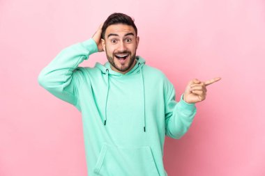 Pembe arka planda izole edilmiş beyaz, yakışıklı genç adam şaşırmış ve parmağıyla kenarı işaret ediyor.
