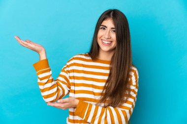 Mavi arka planda izole edilmiş genç beyaz kadın, davet edilmek için ellerini yana uzatıyor.