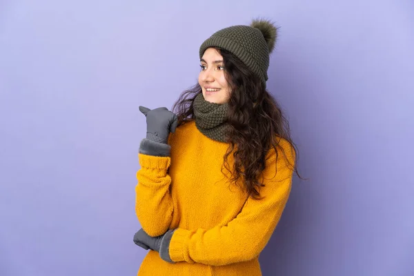 十代のロシアの女の子とともに冬の帽子隔離された紫色の背景に製品を提示する側を指して — ストック写真