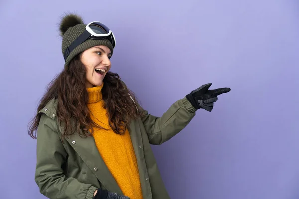 俄罗斯少女 戴着雪板眼镜 与紫色背景隔离 手指指向侧面 并展示了一种产品 — 图库照片
