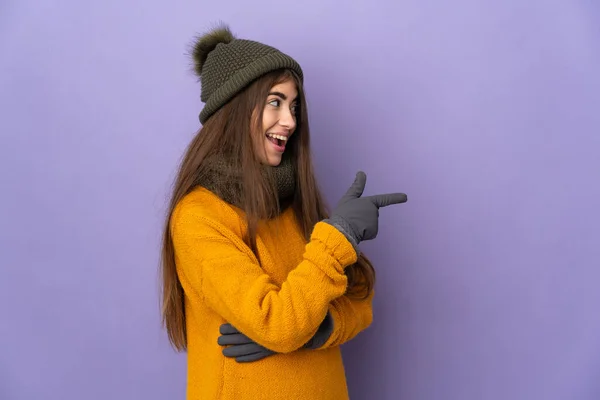 年轻的高加索女孩 戴着冬帽 被紫色背景隔离 手指指向侧面并展示了一种产品 — 图库照片