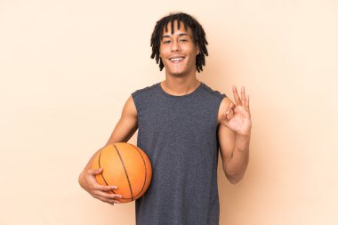 Genç Afrikalı Amerikalı adam bej arka planda izole edilmiş basketbol oynuyor ve OK işareti yapıyor.