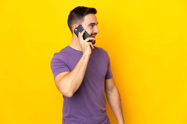 Beyaz tenli yakışıklı adam sarı arka planda izole edilmiş cep telefonuyla sohbet ediyor.