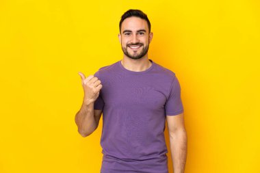 Sarı arka planda izole edilmiş beyaz, yakışıklı genç bir adam bir ürün sunmak için kenarı işaret ediyor.