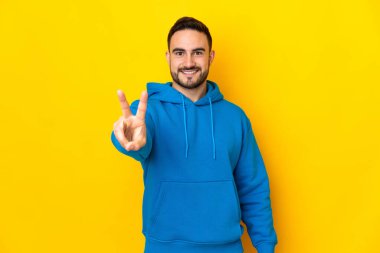 Sarı arka planda izole edilmiş yakışıklı genç adam gülümsüyor ve zafer işareti gösteriyor.