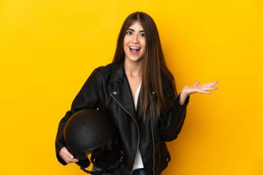 Motosiklet kaskı tutan beyaz bir kadın sarı arka planda izole edilmiş, şok olmuş bir yüz ifadesi var.