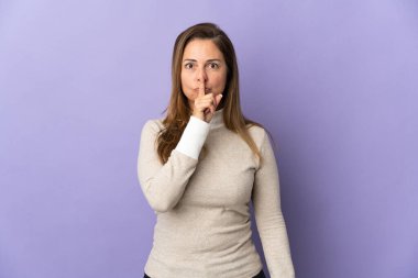 Mor arka planda izole edilmiş orta yaşlı Brezilyalı kadın parmağını ağzına koyan sessizlik işareti gösteriyor.