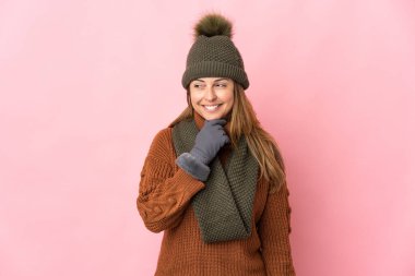 Kış şapkalı orta yaşlı bir kadın pembe arka planda izole edilmiş, yana bakıyor ve gülümsüyor.
