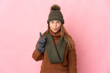 Kış şapkalı orta yaşlı kadın pembe arka planda izole edilmiş. Çözümün ne olduğunu anlamak için parmağını kaldırıyor.