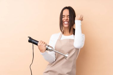 Genç esmer kız el blenderi kullanarak izole bir arka planda zaferi kutluyor.