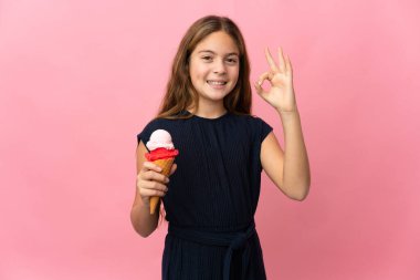 İzole edilmiş pembe arka planda kornet dondurması olan çocuk parmaklarıyla tamam işareti gösteriyor.