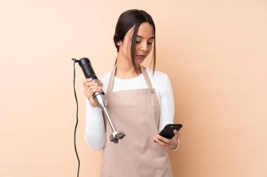 Genç esmer kız el blenderi kullanıyor izole edilmiş arka planda elinde kahve ve cep telefonu var.