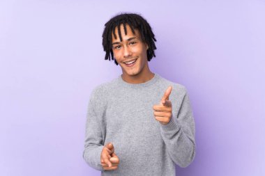 Genç Afro-Amerikalı, mor arka planda izole edilmiş ön tarafı işaret ediyor ve gülümsüyor.