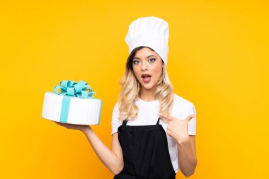 Sarı arka planda büyük bir pasta tutan ergen kız pastası. Şaşırtıcı bir yüz ifadesi var.