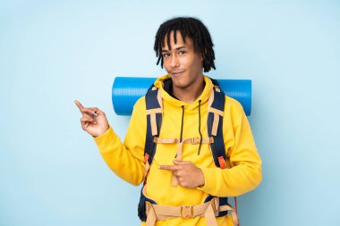 Genç dağcı Afro-Amerikan adam büyük bir sırt çantası ile izole mavi arka planda parmak yan işaret