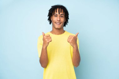 Genç Afrikalı Amerikalı adam mavi arka planda izole edilmiş. Baş parmakları havada ve gülümsüyor.