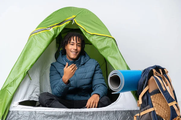 Young african american man inside a camping green tent inviting to come with hand. Happy that you came