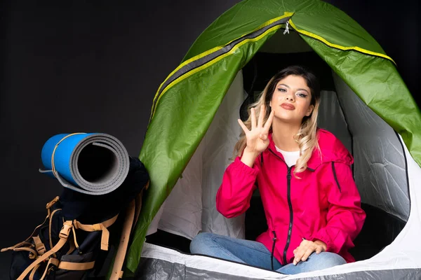 Teenager girl inside a camping green tent isolated on black background happy and counting four with fingers