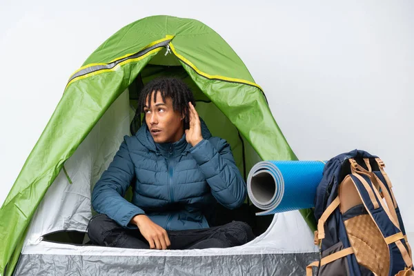 Young african american man inside a camping green tent listening to something by putting hand on the ear
