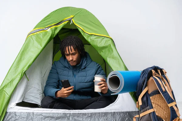 Young african american man inside a camping green tent holding coffee to take away and a mobile