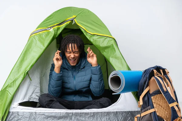 Young african american man inside a camping green tent with fingers crossing